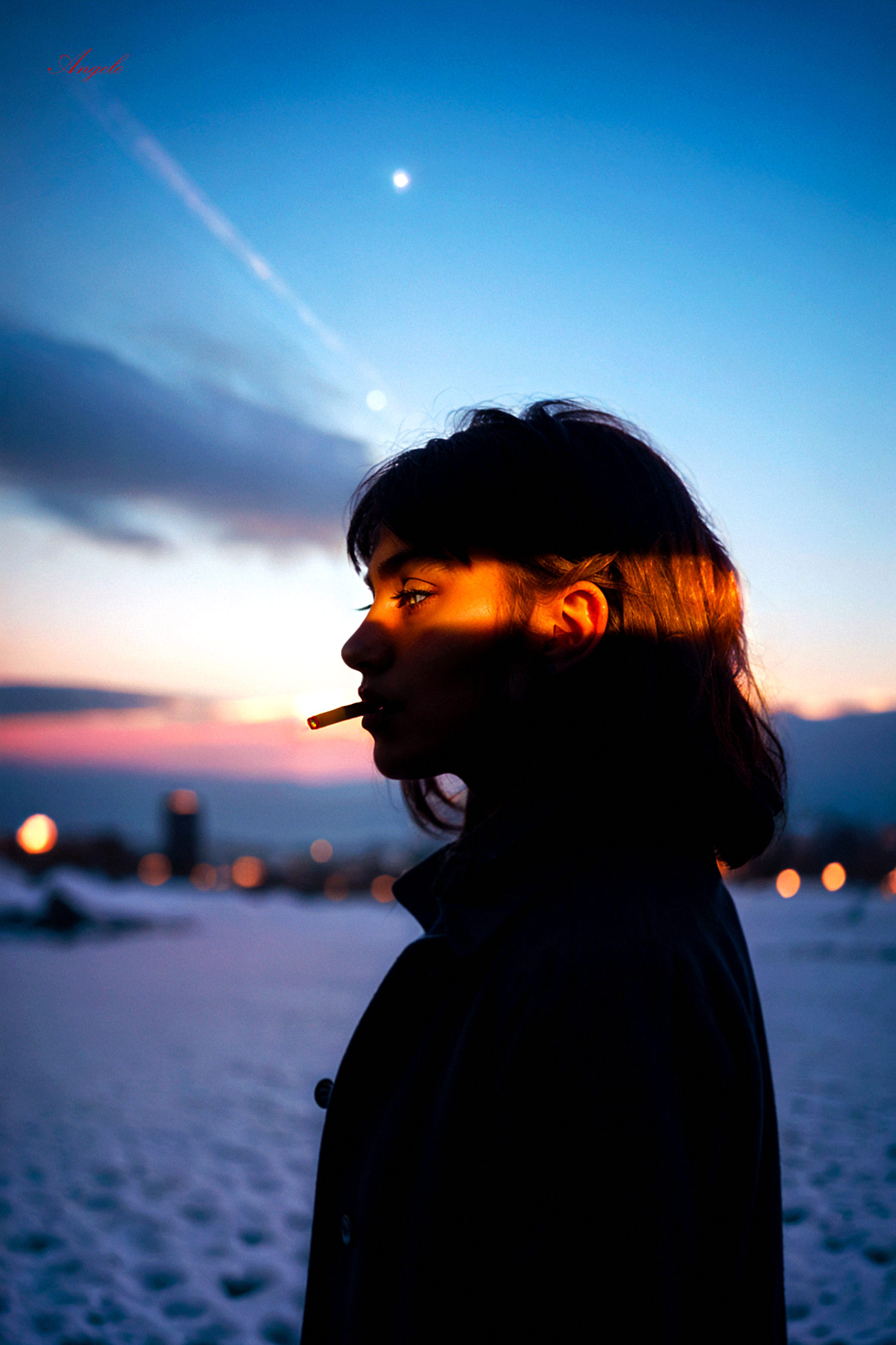 A young woman in profile, facing left, against a stunning twilight sky. The woman has medium-length, dark hair, and she is wearing a dark-colored coat. The background features a snow-covered landscape, with distant city lights and a clear, sunset sky that transitions from deep blue to a warm orange and pink hue near the horizon. The woman holds a lit cigarette in her mouth as a warm, orange ray of sunlight illuminates her face partially, creating an interesting contrast between the cool blue of the sky and the warm tones of light. 