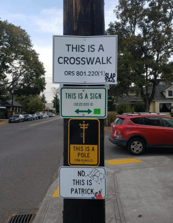 An assortments on signs on a power pole in a city, reading from the top down: "This is a Crosswalk ORS 801.220(1)" "This is a Sign 32.22.020 X" "This is a pole TRN-10.44.11.C)" "No, this is Patrick" the last sign has a photo of Patrick Star from Spongebob Squarepants present on it.